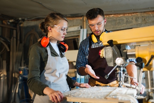 Waist-up portrait of attractive apprentice standing at drill press and listening to her male mentor with attention, he explaining her operating principle of machine